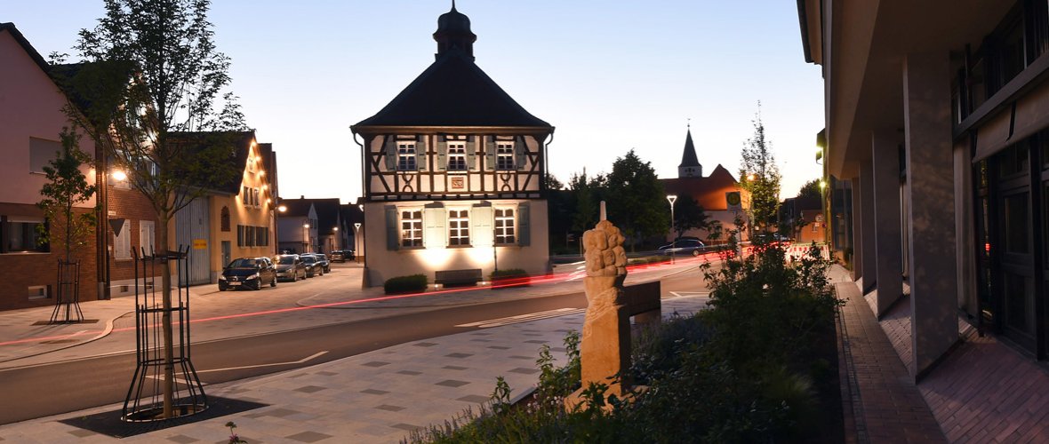 Ansicht auf das beleuchtete Historische Rathaus bei Nacht mit einer  Stehle der Mutterstadter Landfrauen im Vordergrund